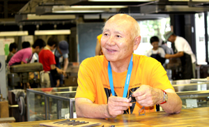 三条鍛冶道場内で話す長谷川館長、後方に見えるのは鍛冶を体験する子どもたち